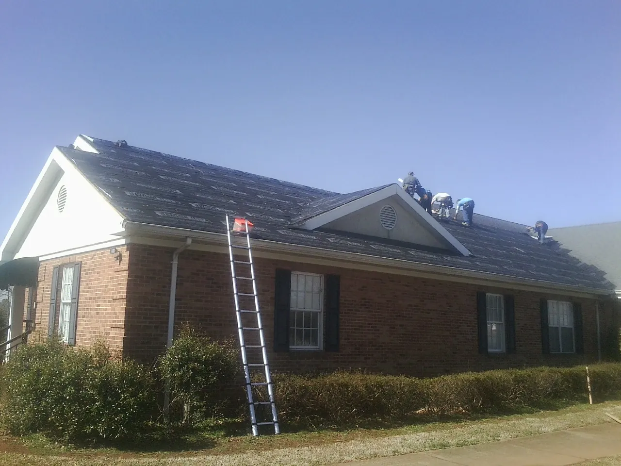 Crew tearing shingles off a brick ranch roof with a ladder set — Metal Roof Repair work by our crew in Scipio Center