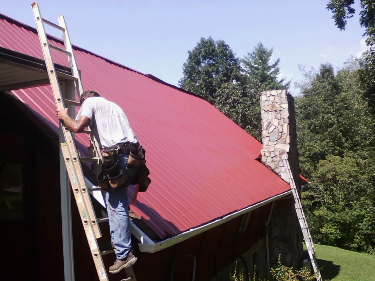 Roofer on a ladder beside a red metal roof and stone chimney — Soffit & Fascia Repair by Roofing Brothers in Scipio Center